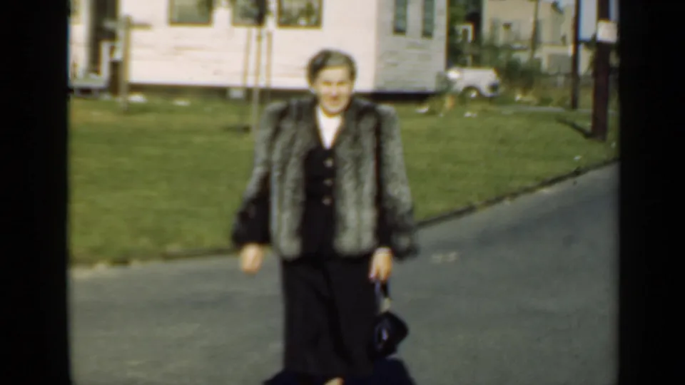 The woman continues toward the camera on a sunny residential street, with houses, lawns, utility poles, and parked cars behind her.