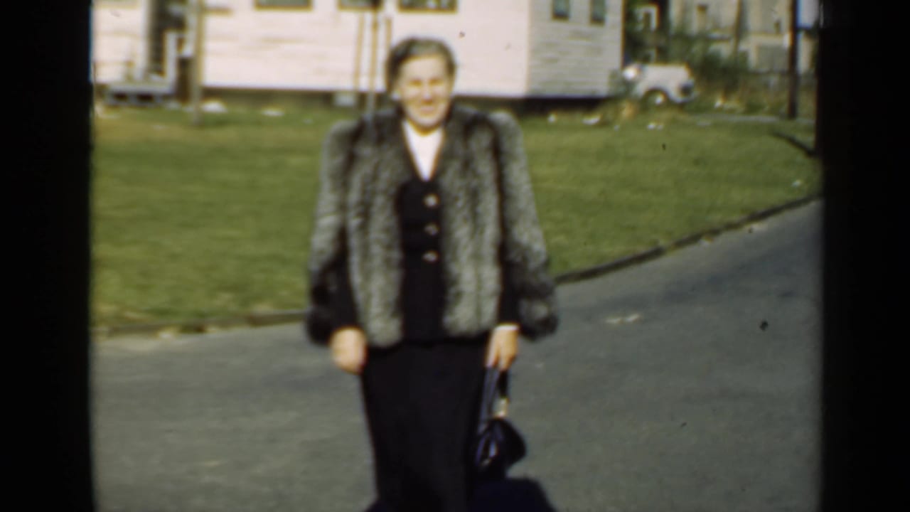 In 1947 New York home-movie footage, an adult woman in a silver-gray fur coat and dark dress walks toward the camera on a residential street.