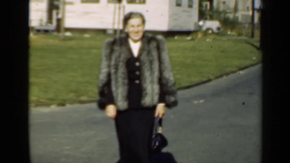 In 1947 New York home-movie footage, an adult woman in a silver-gray fur coat and dark dress walks toward the camera on a residential street.