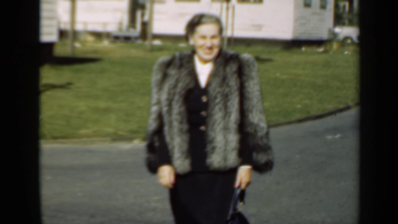 The woman remains centered in the 1947 New York frame, wearing a fur coat over a dark outfit beside a lawn and modest house.