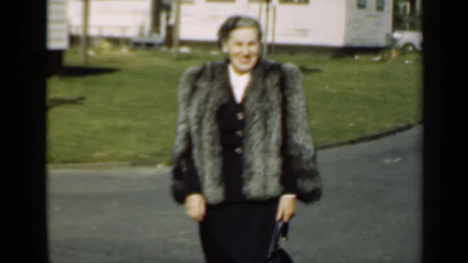 The woman remains centered in the 1947 New York frame, wearing a fur coat over a dark outfit beside a lawn and modest house.