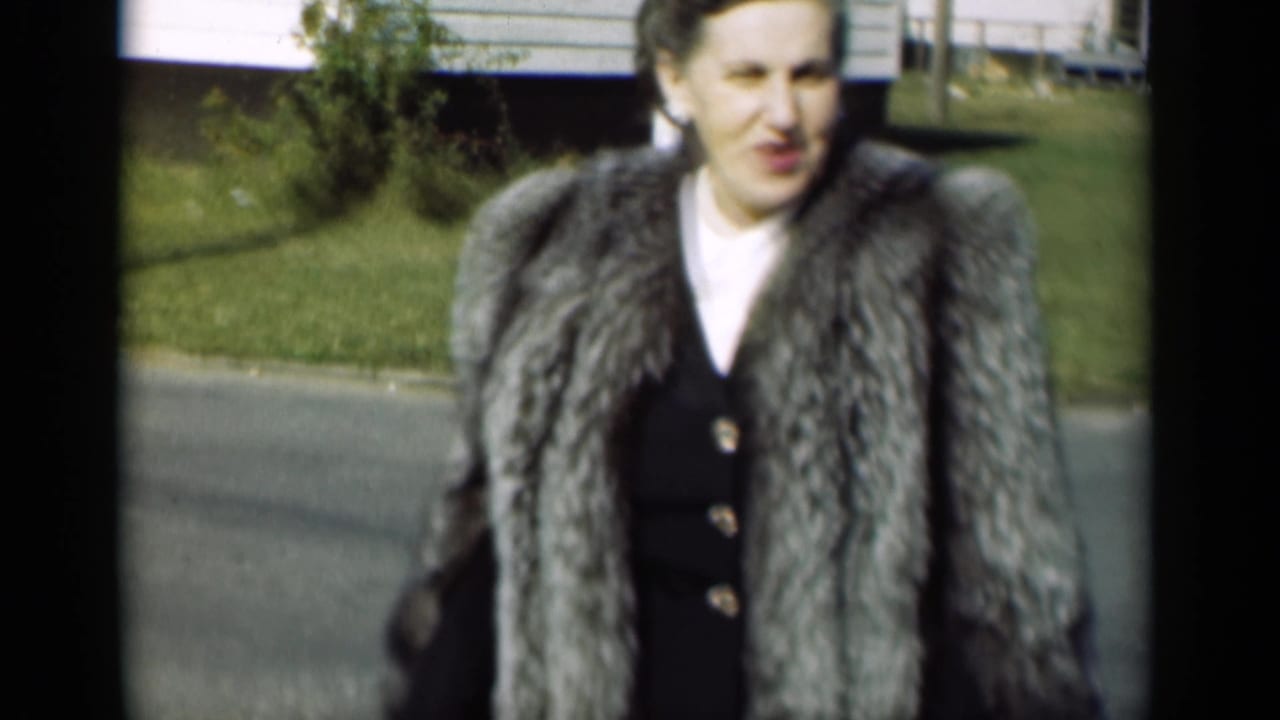 Woman in a silver fur coat walks toward the camera on a sunny residential street.