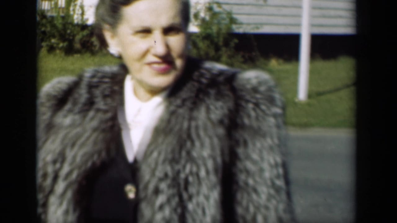 The woman smiles near the camera, with pearl earrings, a fur coat, grass, shrubs, and a house visible behind her.