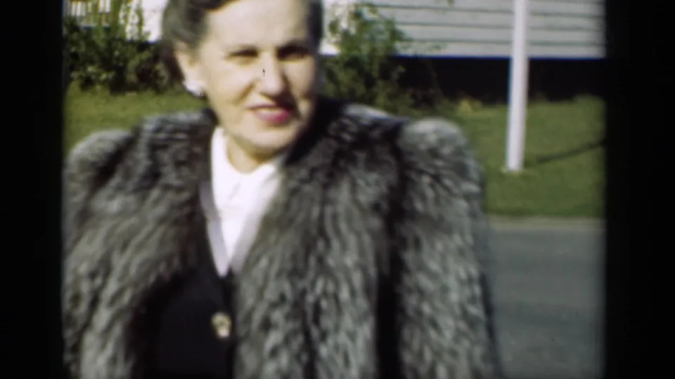 The woman smiles near the camera, with pearl earrings, a fur coat, grass, shrubs, and a house visible behind her.