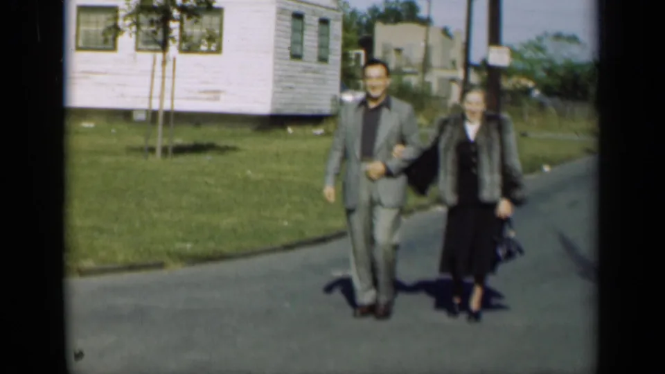 A man in a gray suit and a woman in a fur coat walk arm-in-arm toward the camera on a quiet New York street.