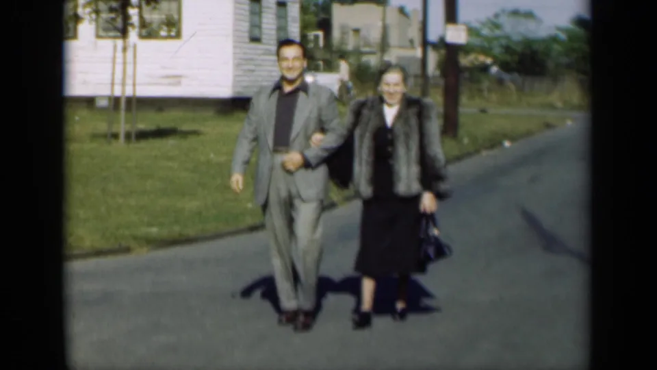 The man and woman continue toward the camera, smiling as they pass houses, grass, and roadside shadows in 1947 New York footage.