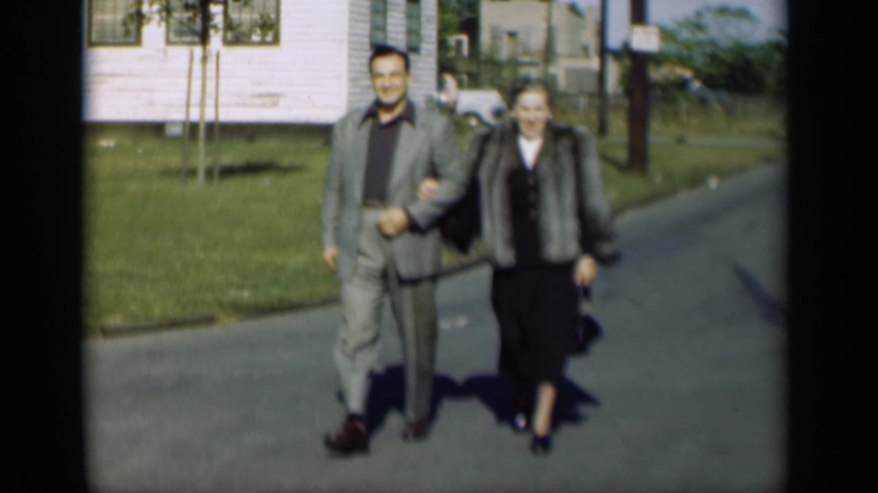 A couple walks arm-in-arm on a paved street with a lawn and white house behind them.