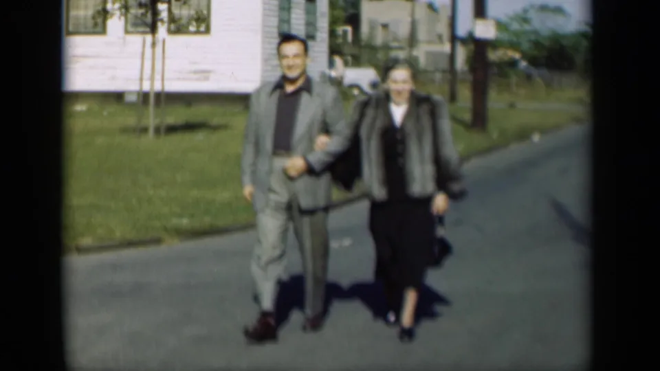 In 1947 New York home-movie footage, a middle-aged couple walks arm-in-arm outdoors near a white house and green lawn.