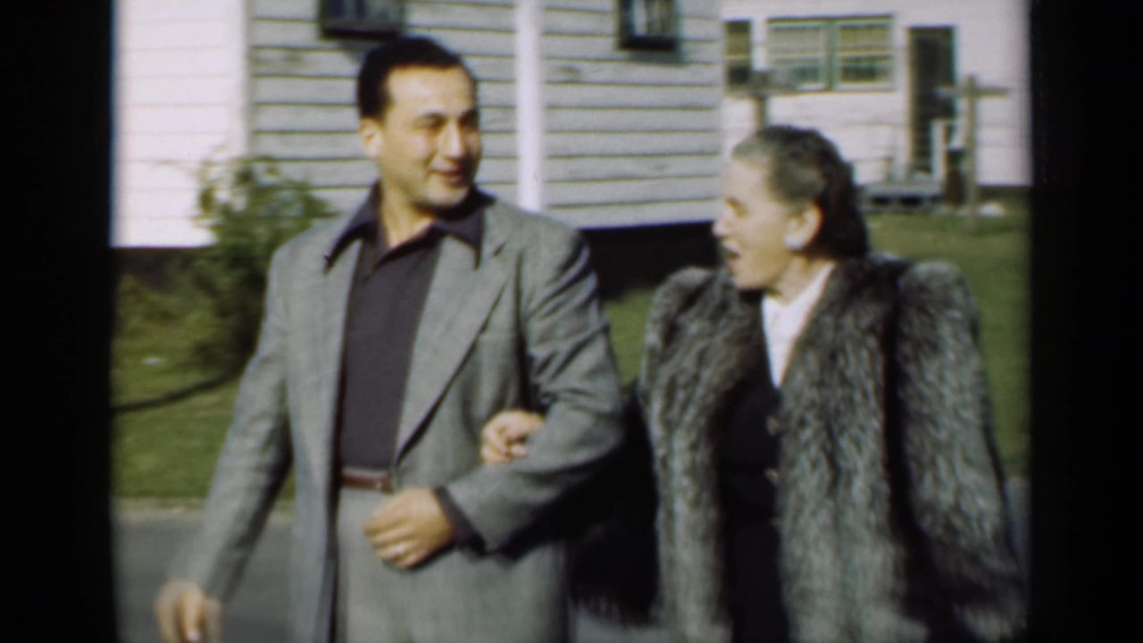 Man in a grey suit walks arm-in-arm with a woman in a fur coat outside a white-sided house.