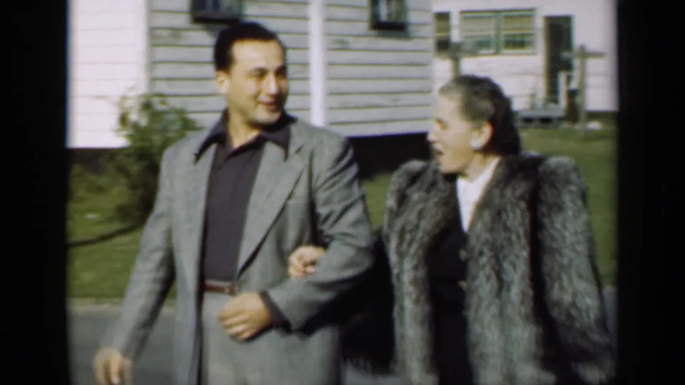 In 1947 New York home-movie footage, a smiling man in a grey suit walks with a woman in a grey fur coat near a white-sided house.