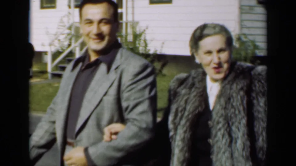 The man in a grey suit and woman in a textured fur coat walk beside a white residential building in bright daylight.