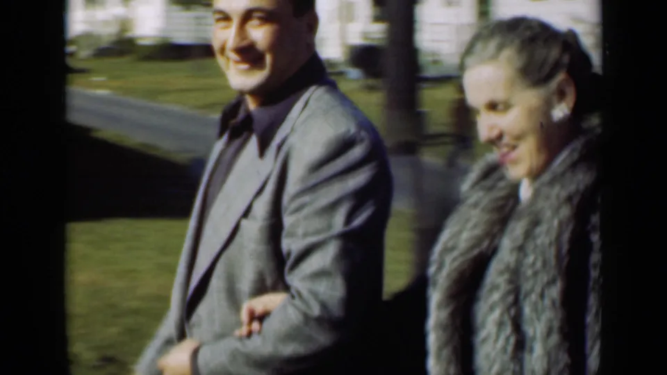 A close 8mm frame shows the couple passing the camera, the woman looking downward and the man smiling in the yard.