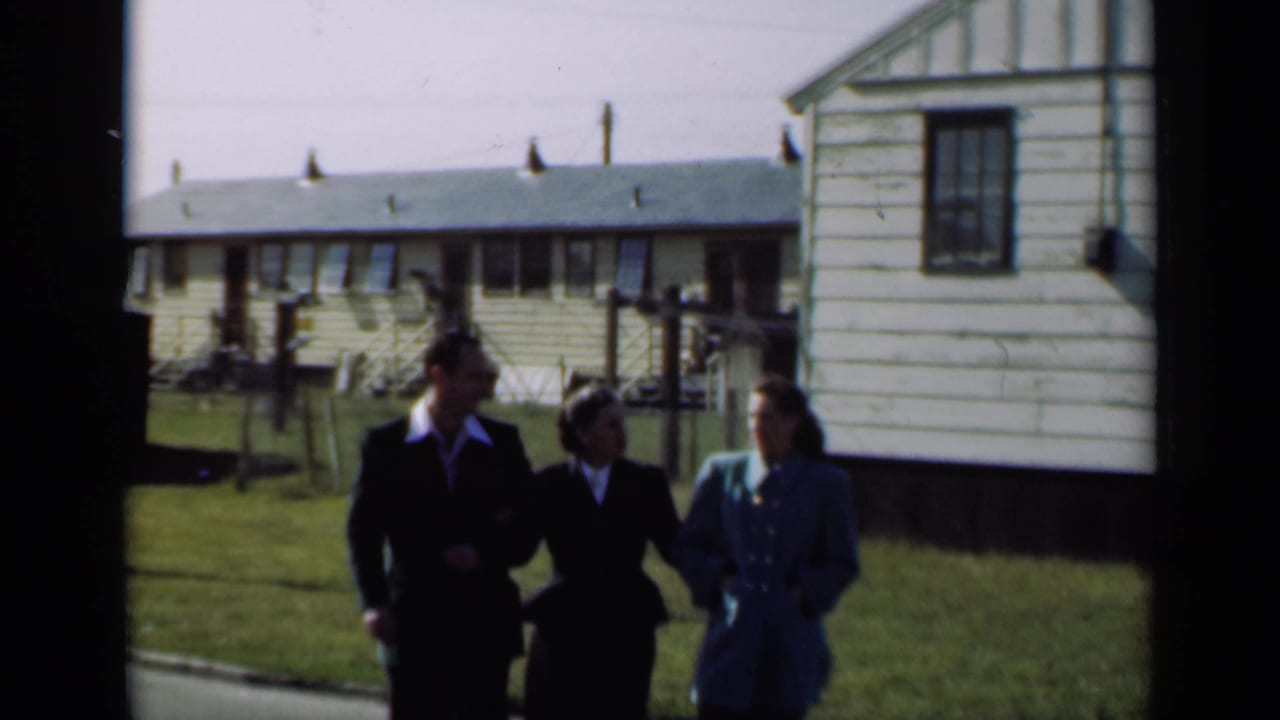 Three dressed adults walk across a lawn near light-colored houses.