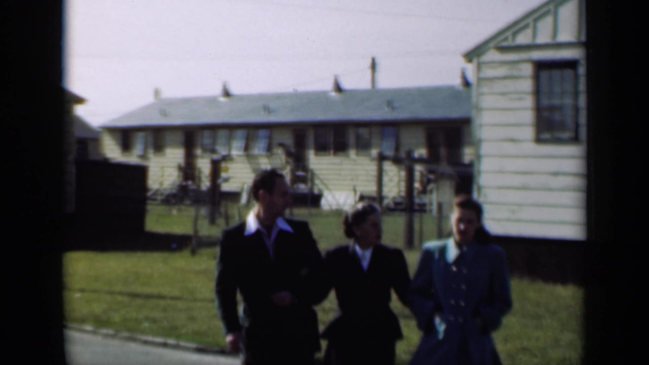 Three adults continue walking toward the camera beside a row of houses.