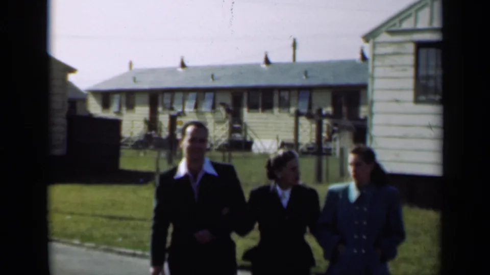 Three dressed adults, including a man and two women, walk arm in arm through a residential lawn in 1947 New York footage.