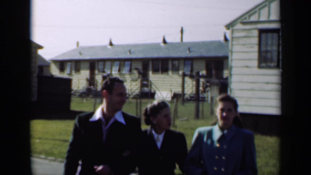 Three adults in 1940s-style dress walk and talk in a New York residential area with light-colored houses behind them.