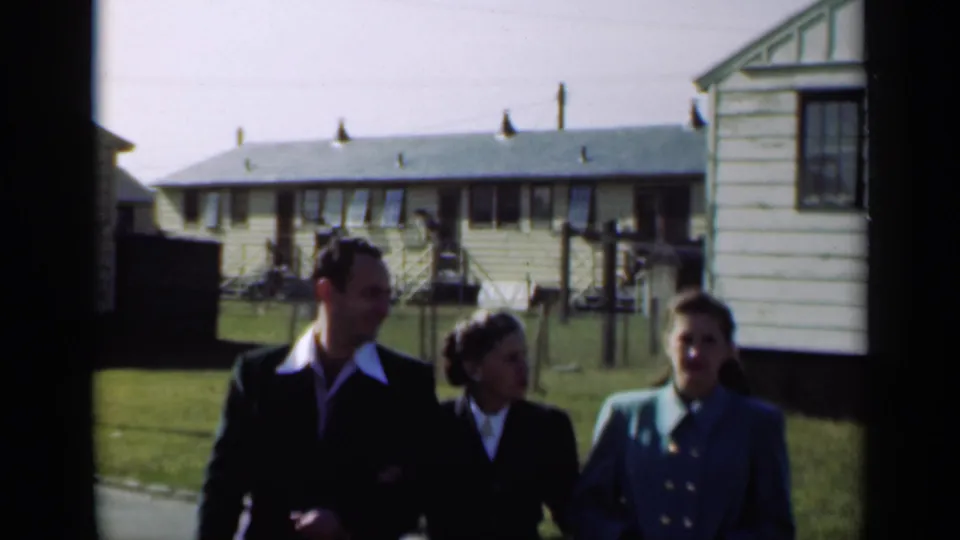 Three adults in 1940s-style dress walk and talk in a New York residential area with light-colored houses behind them.