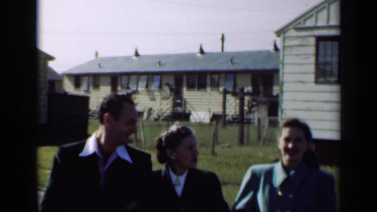 A man and two women walk side by side, smiling and conversing, with small houses and lawns visible in the background.