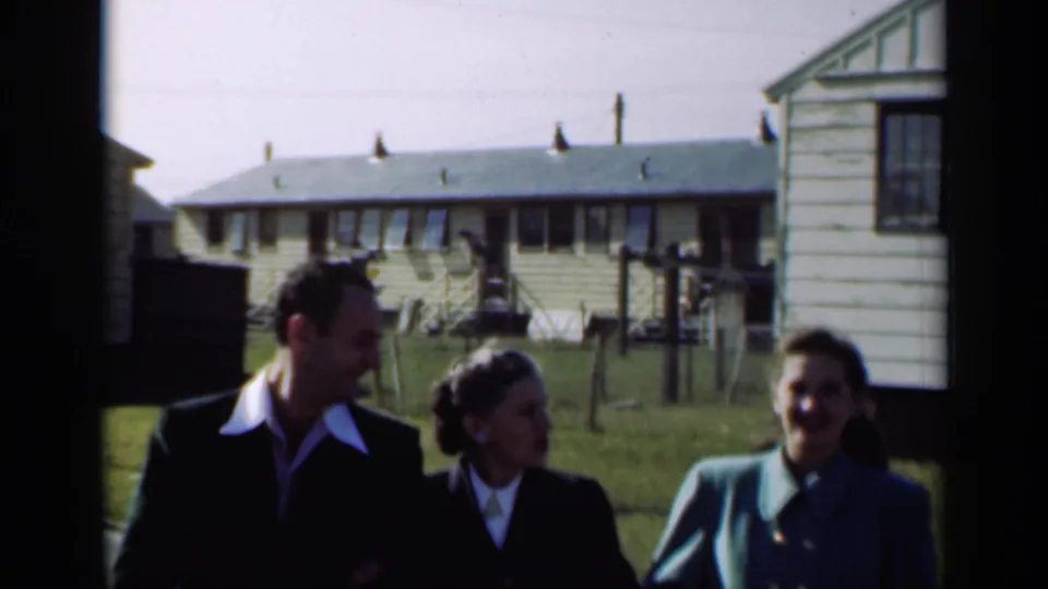A man and two women walk side by side, smiling and conversing, with small houses and lawns visible in the background.