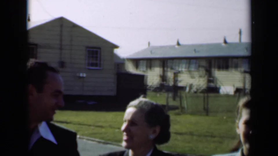 The group continues past the camera in a New York residential setting, with small light-sided houses, grass and fencing behind them.