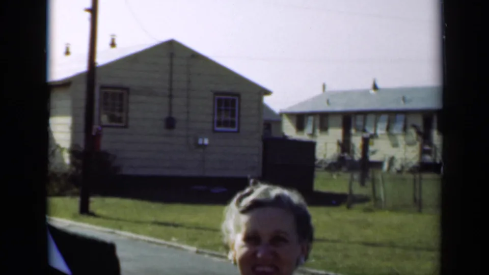 In New York 1947 home-movie footage, an older woman smiles close to the camera on a residential street with modest houses behind her.