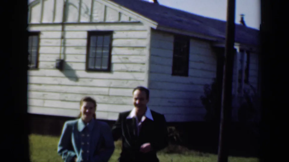 A young couple in New York, 1947, stands outside a modest light-sided suburban house, facing the home-movie camera.