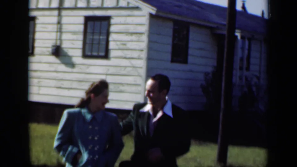 The couple walks together in front of a modest New York suburban house, with the man in a dark suit and the woman in a blue jacket.