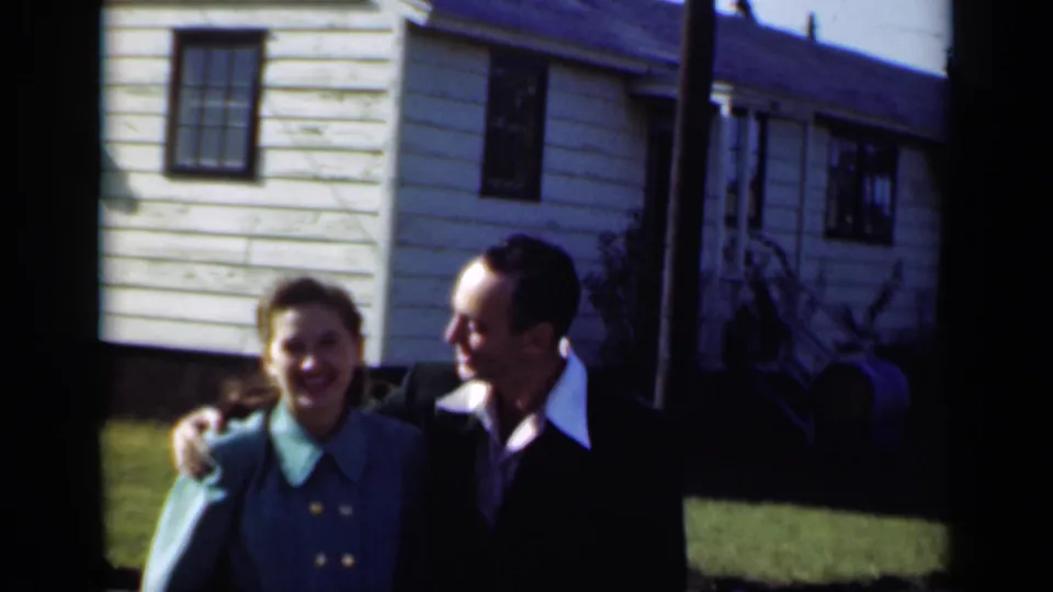 A smiling couple appears arm in arm outside a modest New York house in 1947, captured in soft 8mm color.