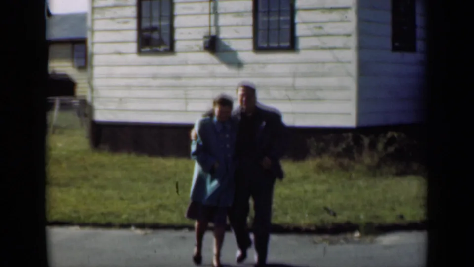 The couple walk arm in arm near a clapboard house and pavement in this 1947 New York home-movie frame.