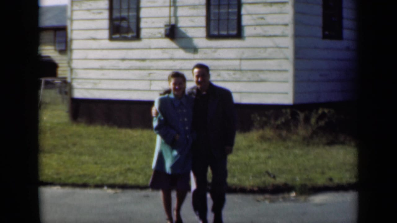 Young couple walking arm-in-arm past a white clapboard house on a sunny street.