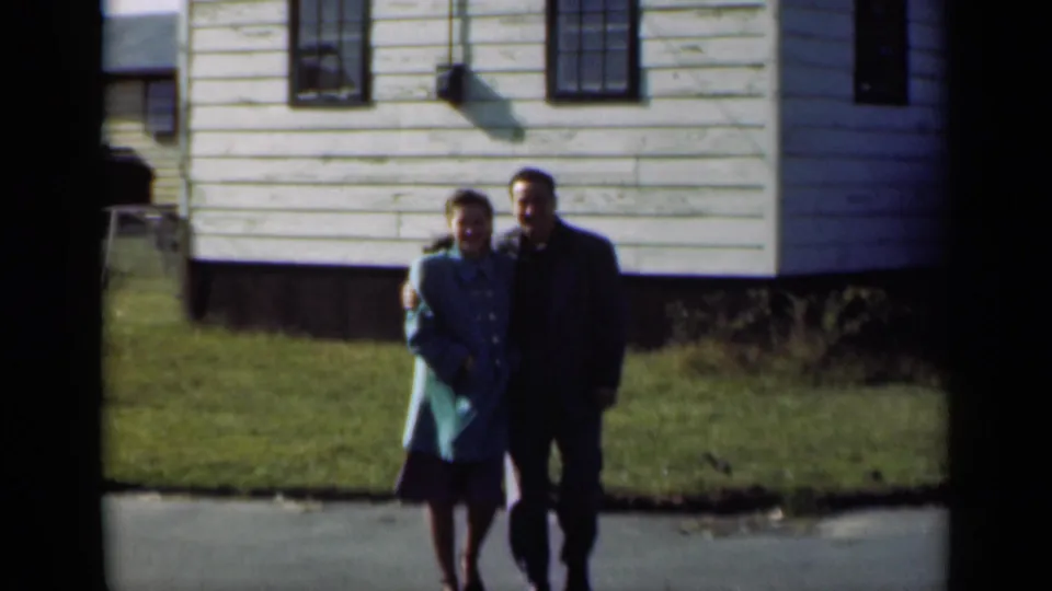 In a 1947 New York home-movie frame, a young couple walks arm-in-arm near a white clapboard house and green lawn.