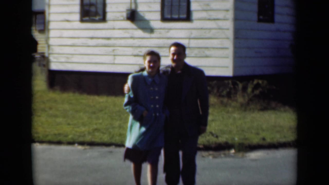 The couple remains arm-in-arm in a 1947 New York residential scene, with a white clapboard house behind them.
