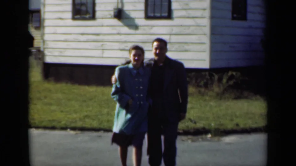 The couple remains arm-in-arm in a 1947 New York residential scene, with a white clapboard house behind them.