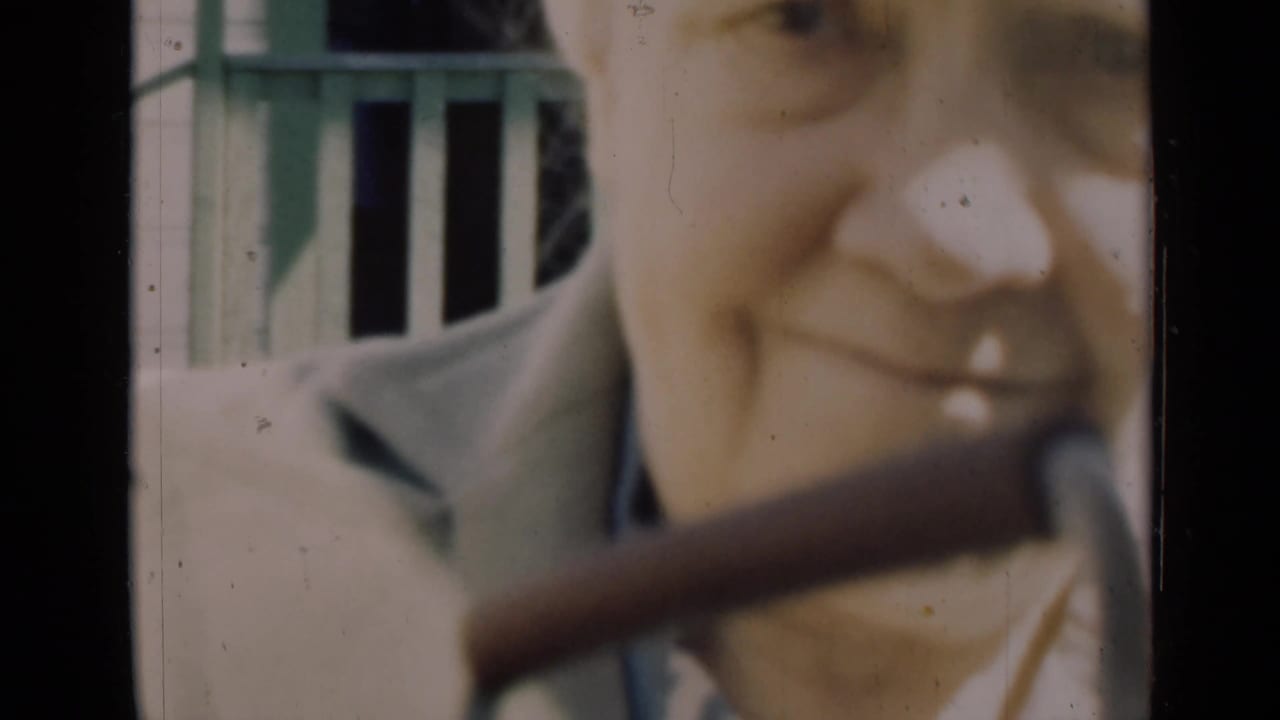 Close 1948 Indiana home-movie frame of an elderly woman smiling near a porch, partly obscured by a pipe or railing.