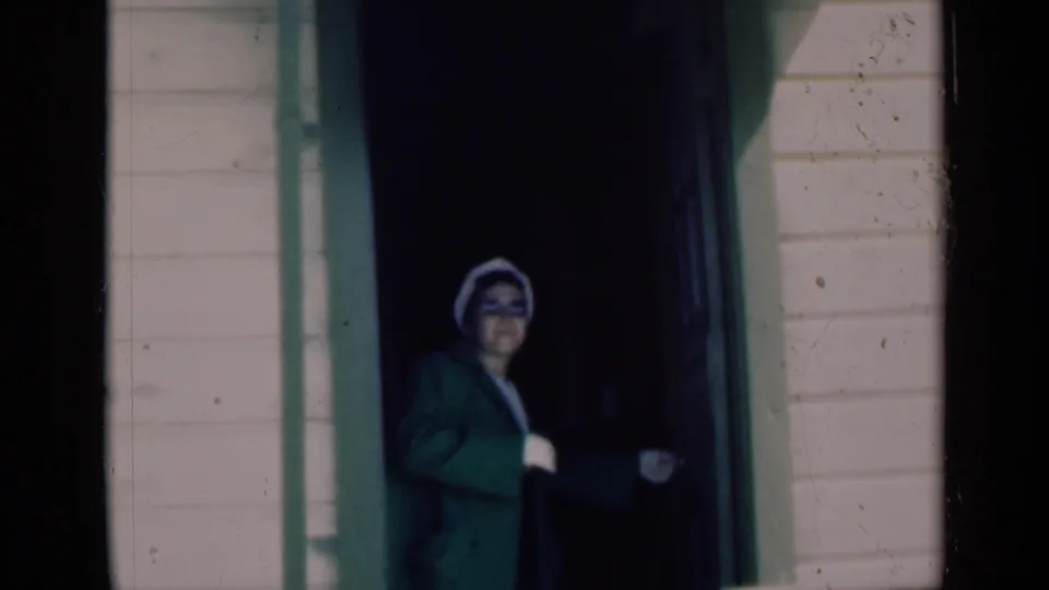 1948 Indiana home-movie frame showing an elderly woman in a green coat and white head covering standing in a dark doorway.
