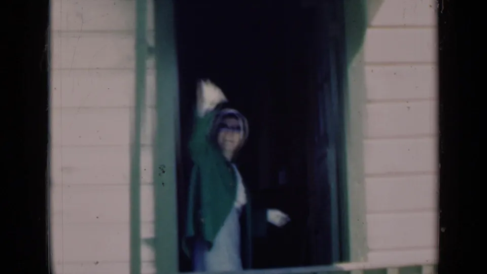 1948 Indiana home-movie frame of an elderly woman in a green coat waving from a dark porch doorway.