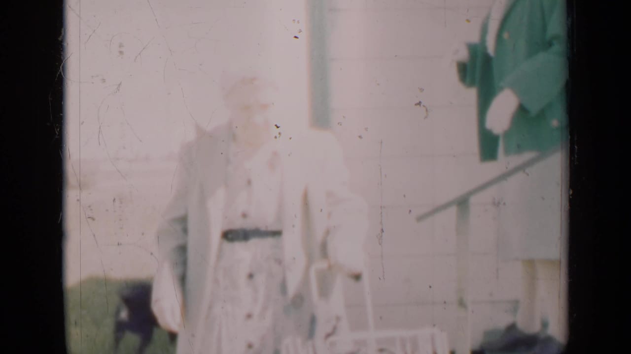 In this 1948 Indiana home-movie frame, a woman in light clothing stands near a porch doorway with a small dog partly visible nearby.