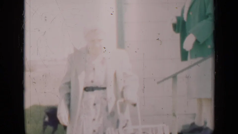 In this 1948 Indiana home-movie frame, a woman in light clothing stands near a porch doorway with a small dog partly visible nearby.