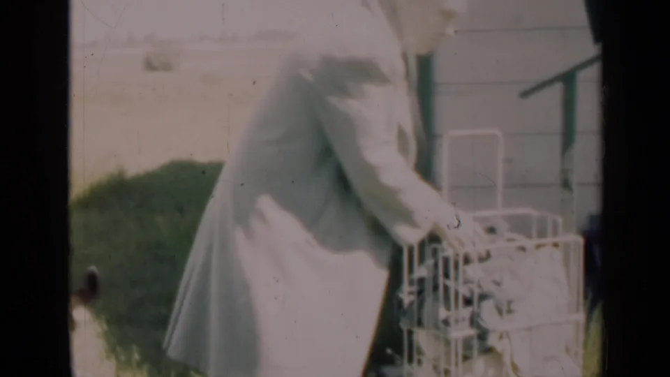 The woman leans over the white carrier beside the porch, with the lawn and house exterior visible in faded 8mm color.