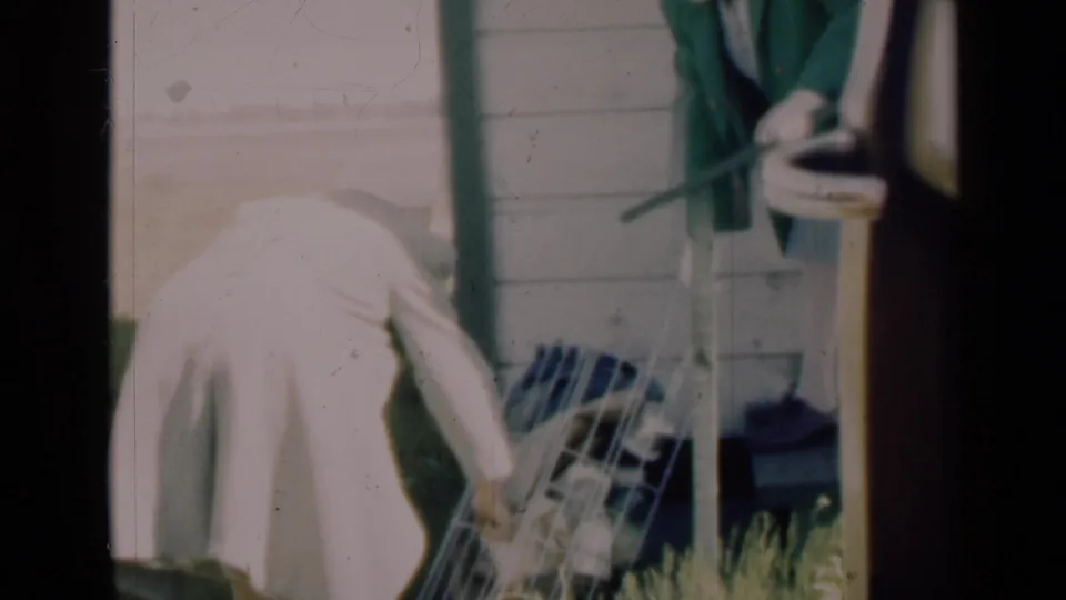 A woman bends to move or lift a tilted white carrier near the porch, while another person remains partly visible by the doorway.