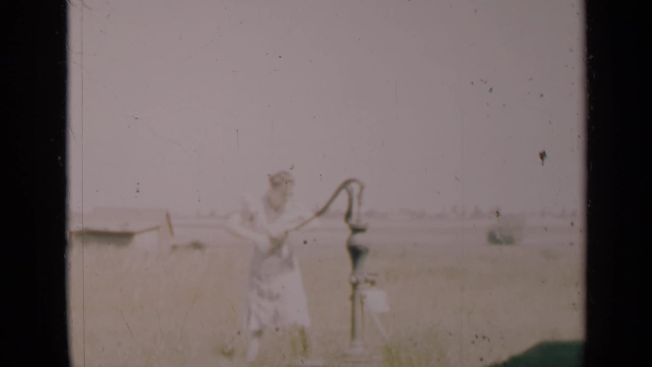 Woman in a light dress stands beside a hand-operated water pump in an open rural field.