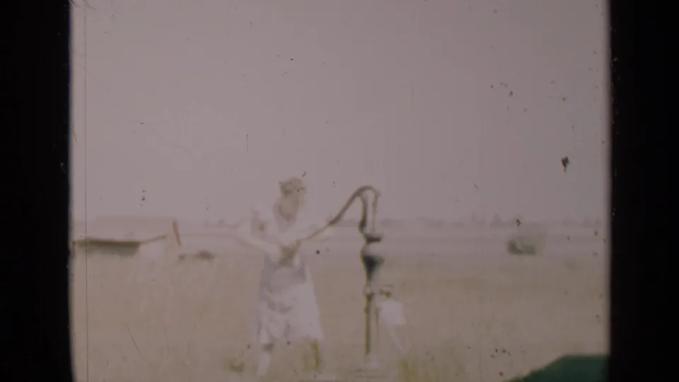 In 1948 Indiana home-movie footage, a woman in a light dress appears beside a metal hand pump in a faded rural field scene.