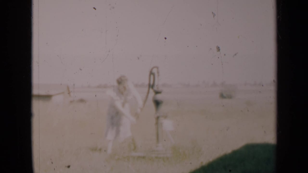 Woman works the hand pump with a rural field and small distant structure behind her.