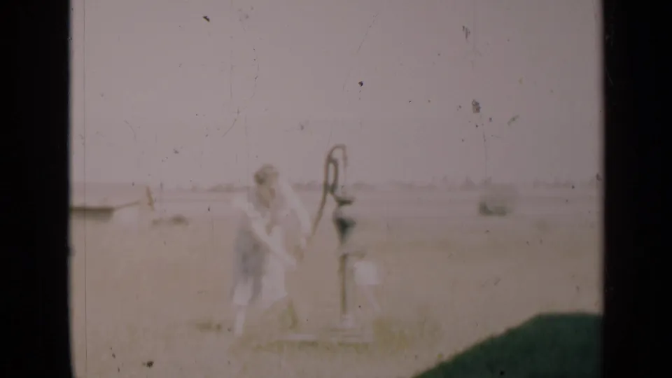 The woman continues pumping water at a metal hand pump in rural Indiana, with a pale sky and low background structure visible.
