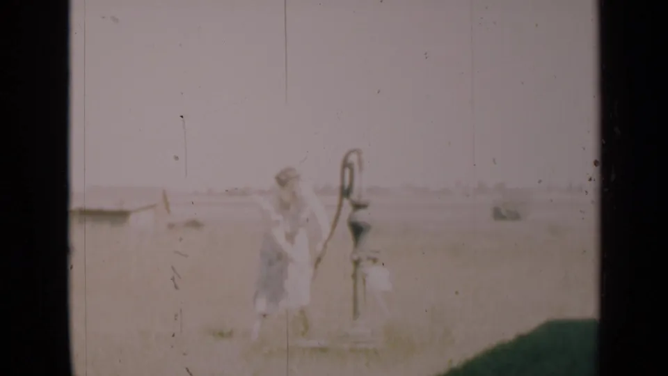 A soft 8mm frame shows the woman near the hand pump, surrounded by open rural ground and a washed-out horizon.