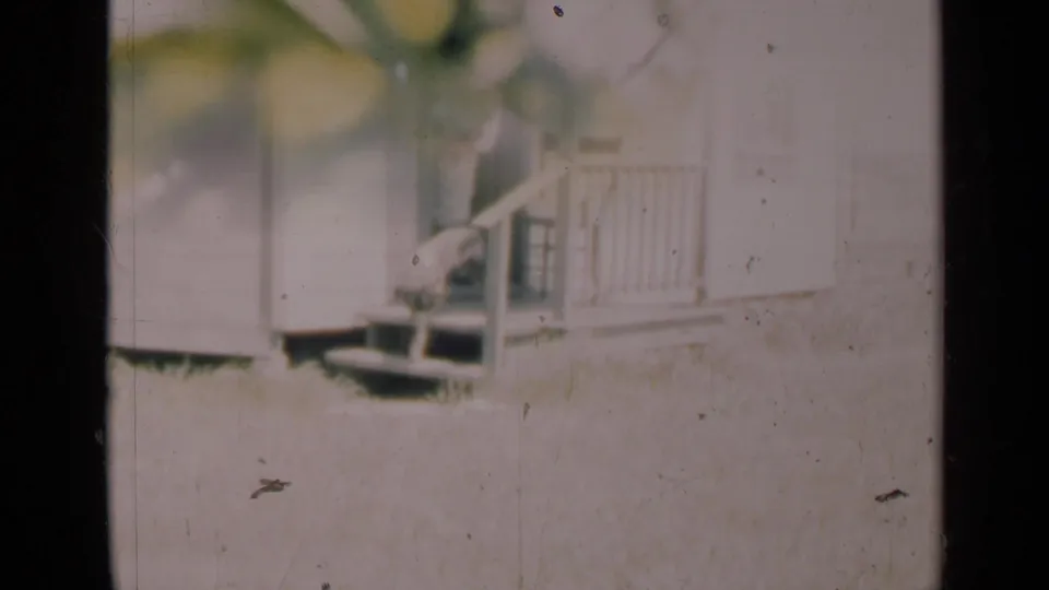 A distant person stands or sits near a white house porch with a dog nearby in the supplied 1948 Indiana home-movie clip.