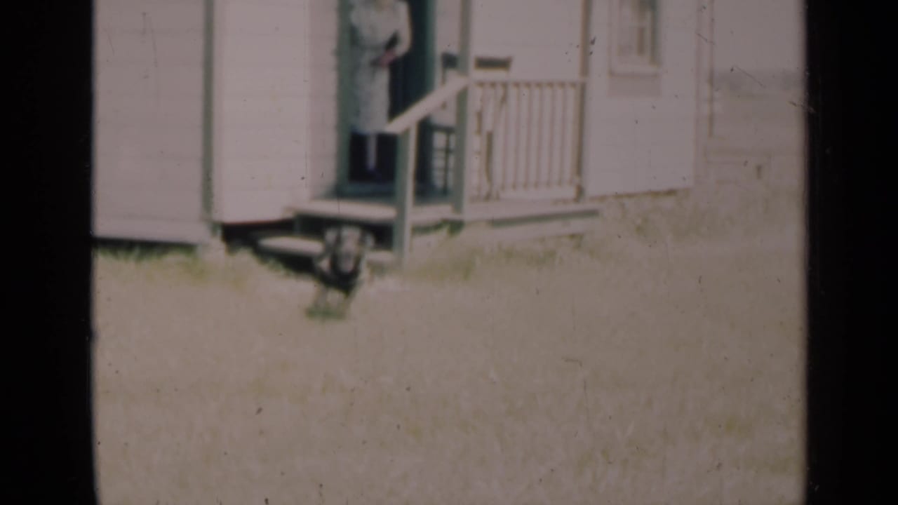 In a 1948 Indiana 8mm home-movie frame, a woman stands at a farmhouse doorway near a porch railing and grassy yard.