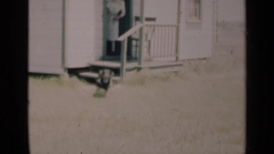 In a 1948 Indiana 8mm home-movie frame, a woman stands at a farmhouse doorway near a porch railing and grassy yard.