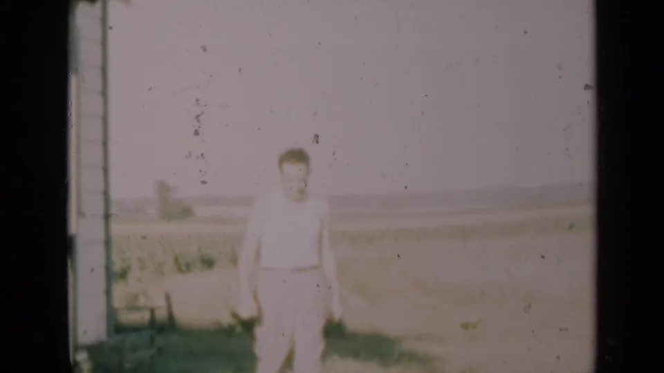 A man in light clothing walks near a rural house with an open field and distant horizon behind him, consistent with the 1948 Indiana setting.