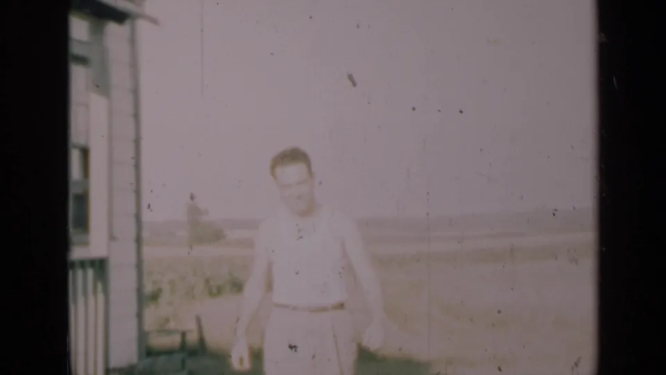 A man walks beside a farmhouse exterior, with an open rural field and soft horizon visible in the faded 8mm frame.